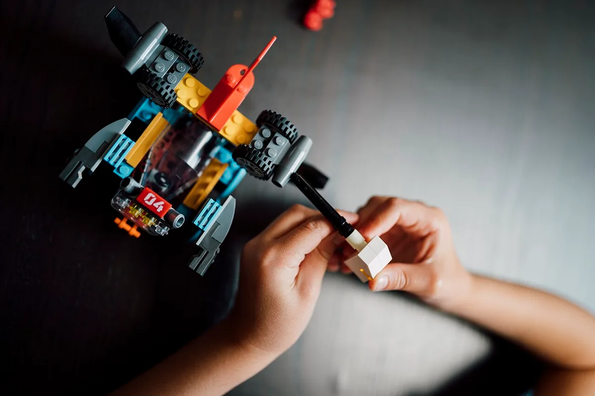Kinderhanden die een klein wit blokje met een zwarte antenne in elkaar zetten, met daarnaast een gebouwd voertuig van gekleurde bouwsteentjes op een donkere tafel